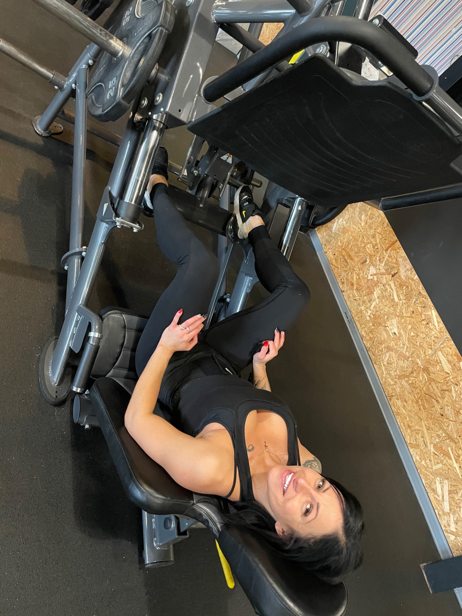 Kobieta w czarnym stroju sportowym ćwiczy na maszynie do wyciskania nogami na siłowni, uśmiechając się do kamery, z widocznymi ciężarami i drewnianym panelem ściennym w tle.
