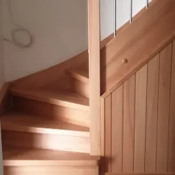 Kątowy widok na jasne, drewniane schody z metalowymi poręczami, widoczne drewniane wykończenie ściany w pionowe panele i fragment brązowej podłogi.