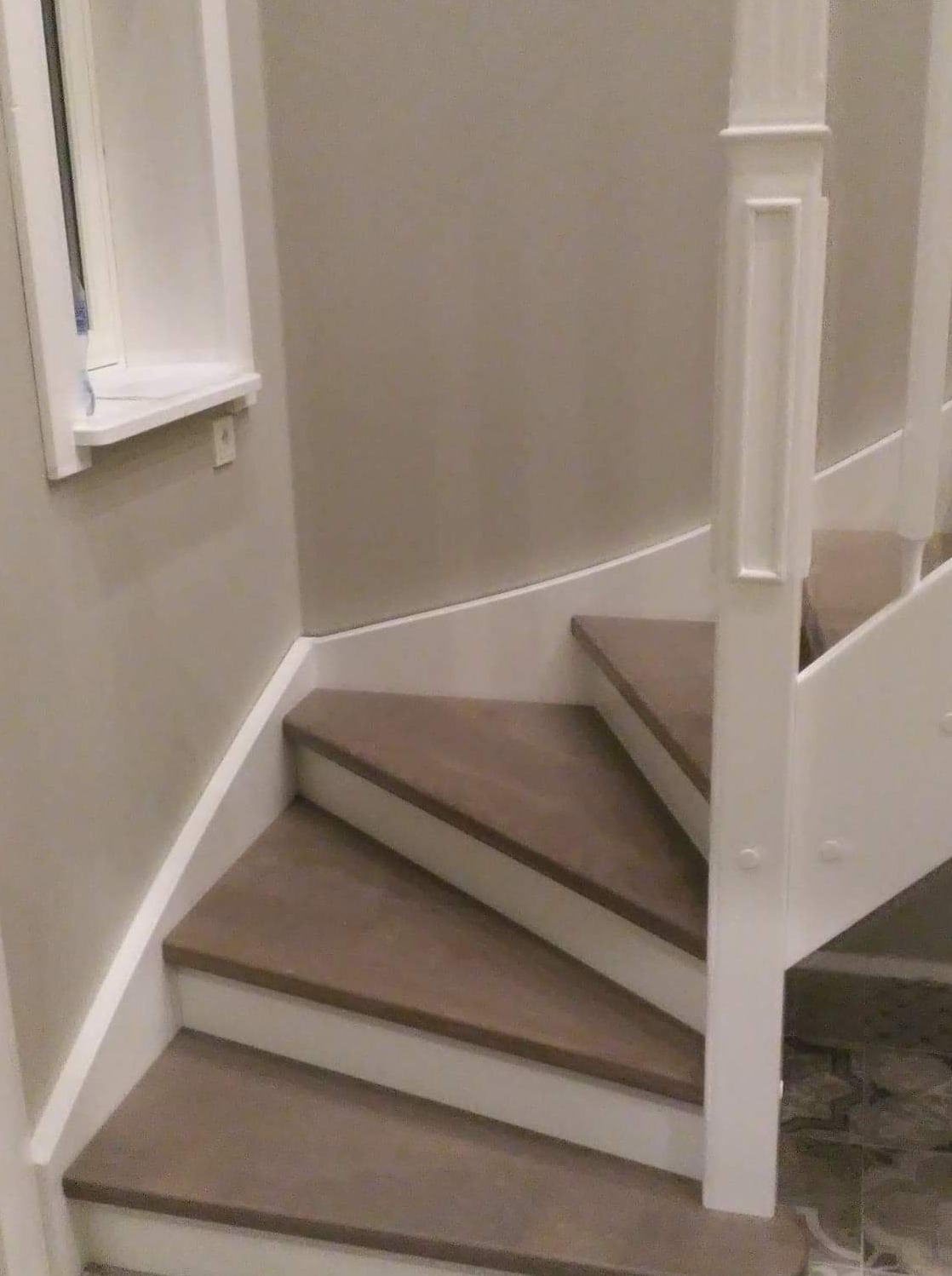 Zakręcone schody z drewnianymi stopniami w odcieniach brązu, białe barierki i listwy przypodłogowe, widoczne gniazdko elektryczne na ścianie.