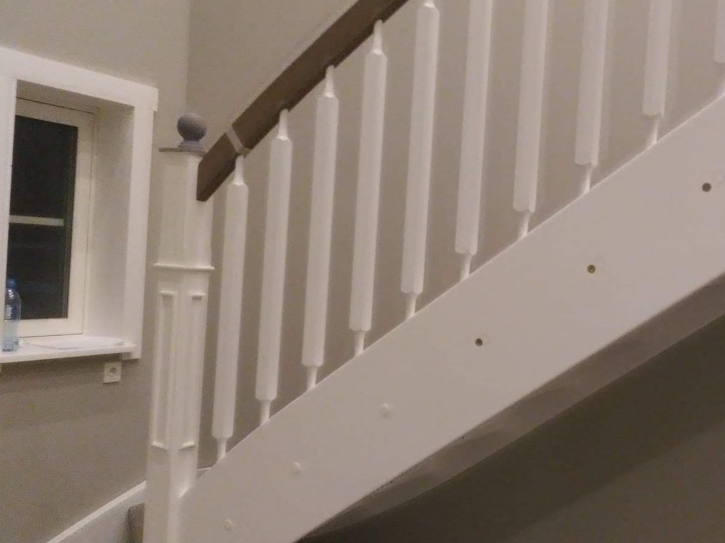 Fragment białych schodów z drewnianą poręczą, widoczne szpachlowane otwory po wkrętach i fragment okna z butelką na parapecie.