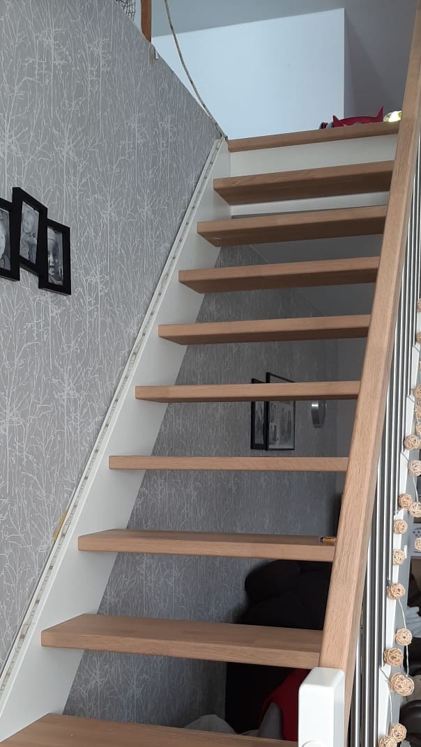 Drewniane schody z białymi bokami i poręczą, oświetlenie LED przy ścianie, szara tapeta w tle.