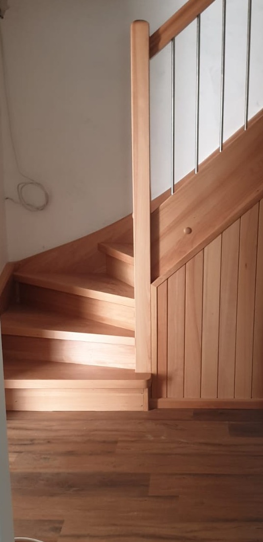 Kątowy widok na jasne, drewniane schody z metalowymi poręczami, widoczne drewniane wykończenie ściany w pionowe panele i fragment brązowej podłogi.