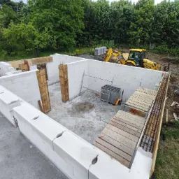 Budowa domu w Andrychowie: widok z góry na fundamenty i ściany z bloczków, zbrojenie na obrzeżach, żółta koparka w tle, teren budowy w otoczeniu zieleni.