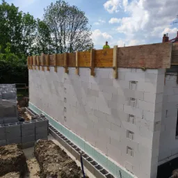 Wznoszenie ściany z białych bloczków, widoczne bloczki fundamentowe z hydroizolacją oraz drewniane szalunki z wystającą armaturą, w tle pracownik w żółtej kamizelce.
