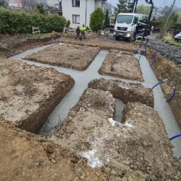 Wylane betonowe fundamenty pod budynek jednorodzinny, widoczny zbrojony szkielet w wykopach, w tle dom i betoniarka z pompą. Na krawędzi wykopu leżą niebieskie rury.