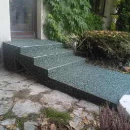 Zewnętrzne schody wejściowe z posadzką żywiczną w kolorze zielonym, wykończone metalowymi listwami, prowadzące do drzwi wejściowych, otoczone roślinnością.