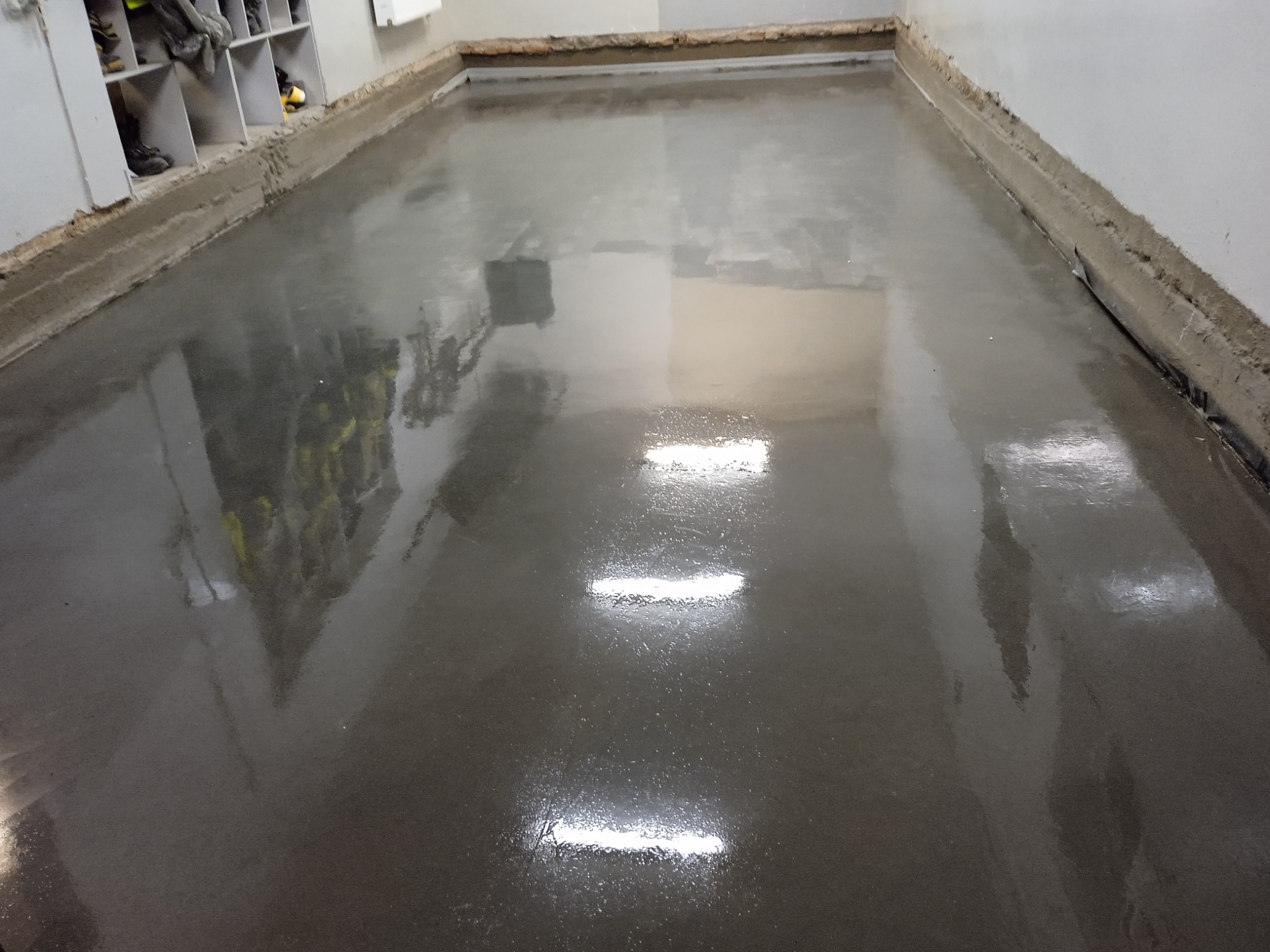 Świeżo wylana, błyszcząca, szara posadzka w pomieszczeniu z szarą ścianą i regałem na buty w tle.