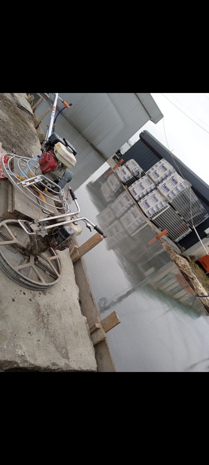 Zacieraczka mechaniczna obok świeżo wylanej, gładkiej posadzki jastrychowej, z widocznymi deskami szalunkowymi i paletami materiałów budowlanych w tle.