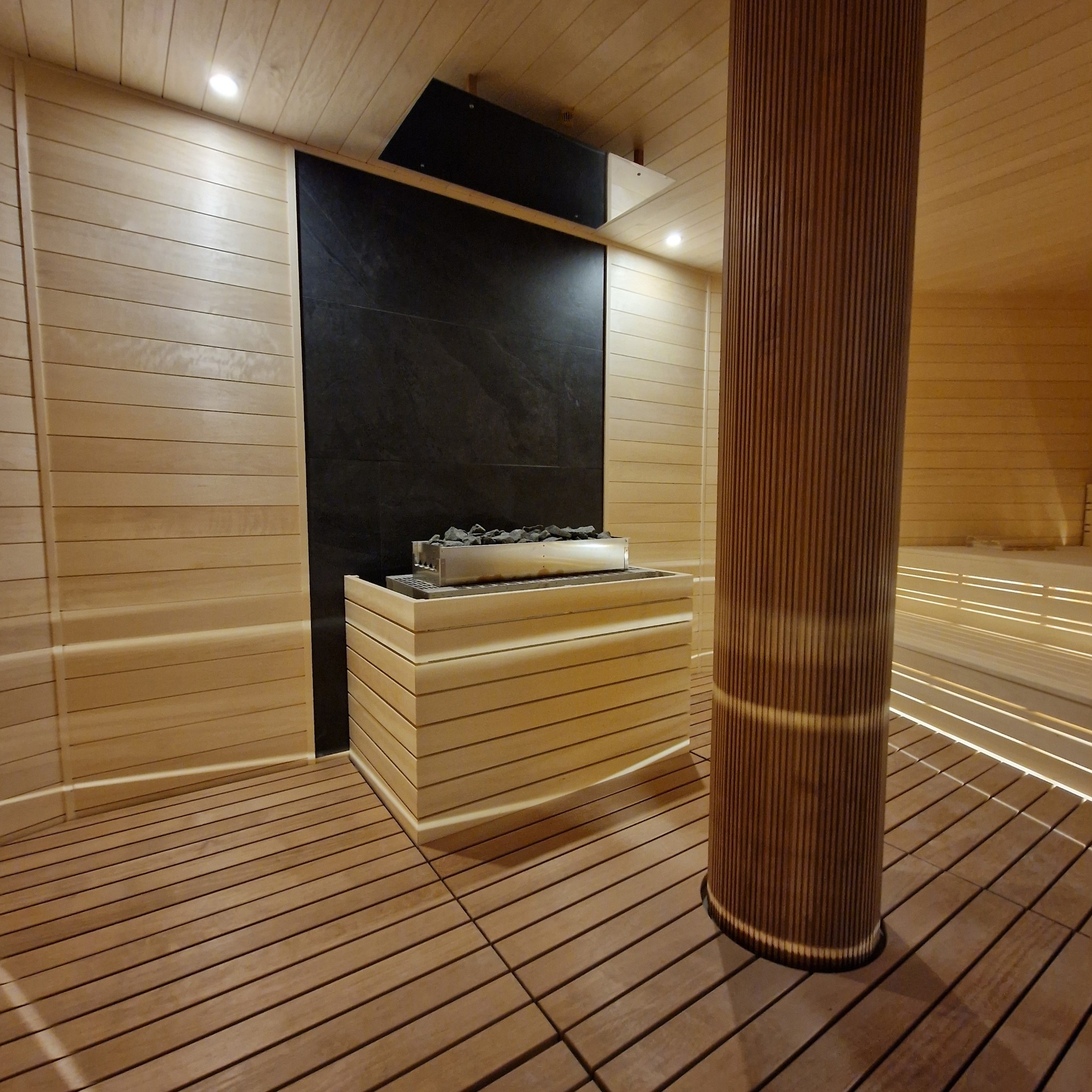 Wnętrze sauny z drewnianym wykończeniem, piecem z kamieniami i oświetleniem pod ławkami. Kolumna z drewna dodaje unikalnego charakteru.