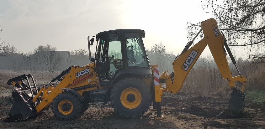 Żółto-czarna koparko-ładowarka JCB na ziemi, z widocznymi podporami stabilizującymi i łyżką kopiącą, w tle drzewa i budynek w porannym świetle.