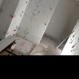 Ściany w trakcie układania białych płytek imitujących marmur, z widocznymi krzyżykami dystansowymi i przewodami pod instalację elektryczną; na podłodze kartony z płytkami GRES i narzędzia glazurnicze.