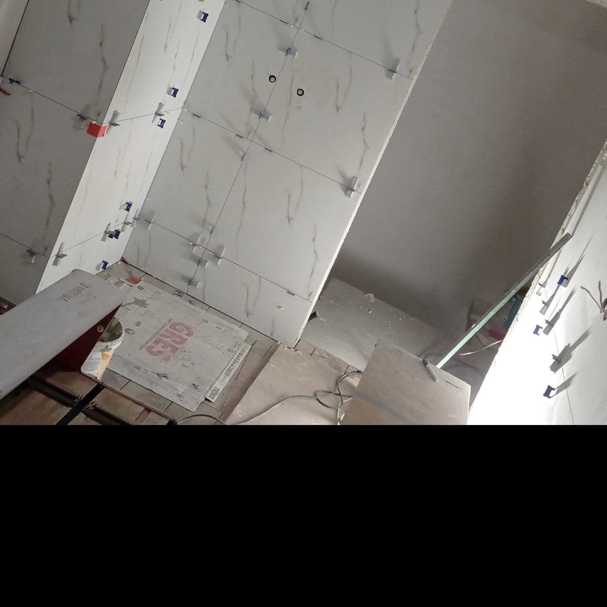 Ściany w trakcie układania białych płytek imitujących marmur, z widocznymi krzyżykami dystansowymi i przewodami pod instalację elektryczną; na podłodze kartony z płytkami GRES i narzędzia glazurnicze.