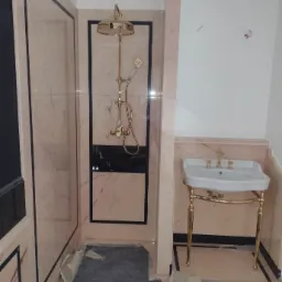 Łazienka w trakcie wykańczania: złota armatura prysznicowa i umywalka na złotym stelażu, marmurowe płytki z czarnymi obramowaniami, zabezpieczone podłogi.