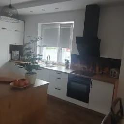 Jasna kuchnia z wyspą, stalową lodówką, czarnym okapem i dekoracyjnym panelem nad płytą grzewczą.