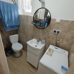 Fanabis Łukasz Biedziński - Ustawna łazienka z beżowymi płytkami, białą armaturą i pralką Candy, widok na okrągłe lustro z półką i wiszący niebieski ręcznik.