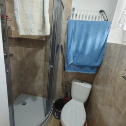 Fanabis Łukasz Biedziński - Narożna kabina prysznicowa z jasną zasłoną, biała toaleta z niebieskim ręcznikiem na grzejniku drabinkowym, beżowe płytki na ścianach, wiadro z mopem obok toalety.