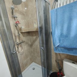 Fanabis Łukasz Biedziński - Narożna kabina prysznicowa z beżowymi płytkami, widoczny zestaw prysznicowy z półką na kosmetyki, niebieski ręcznik na grzejniku, wiadro z mopem obok toalety.