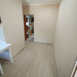 Fanabis Łukasz Biedziński - Wnętrze po remoncie: beżowe ściany, podłoga wyłożona podłużnymi płytkami imitującymi drewno, widok na sąsiednie pomieszczenie z meblami.