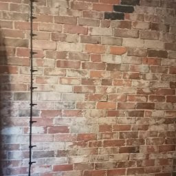 Fanabis Łukasz Biedziński - Ściana z cegły dekoracyjnej w odcieniach czerwieni, brązu i szarości z pionowo poprowadzonym kablem elektrycznym mocowanym za pomocą uchwytów.