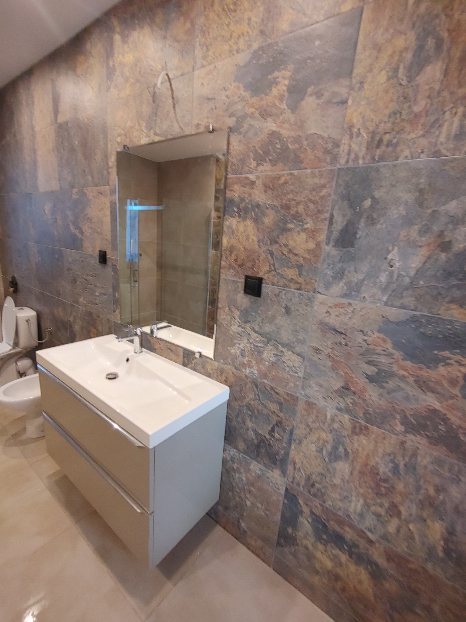 Ukończona łazienka z umywalką nablatową na szafce z szufladami, lustrem bez ramy i toaletą w tle; ściany wykończone płytkami imitującymi kamień w odcieniach brązu i szarości.