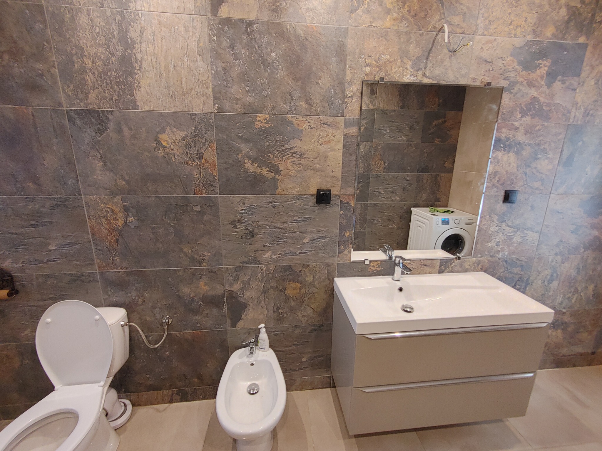 Ukończona łazienka z płytkami imitującymi kamień, białą umywalką z szafką, bidetem i toaletą; w lustrze odbija się pralka.