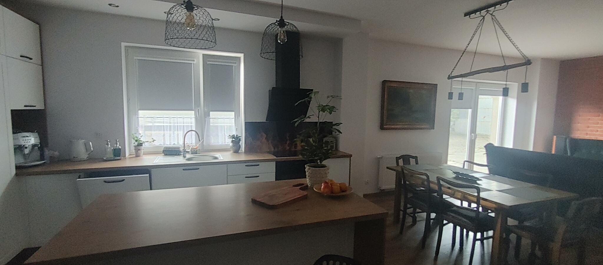 Jasna kuchnia z wyspą, widok na stół jadalny i kominek z roślinami, oświetlona lampami w stylu industrialnym, z drewnianym blatem i dodatkami.