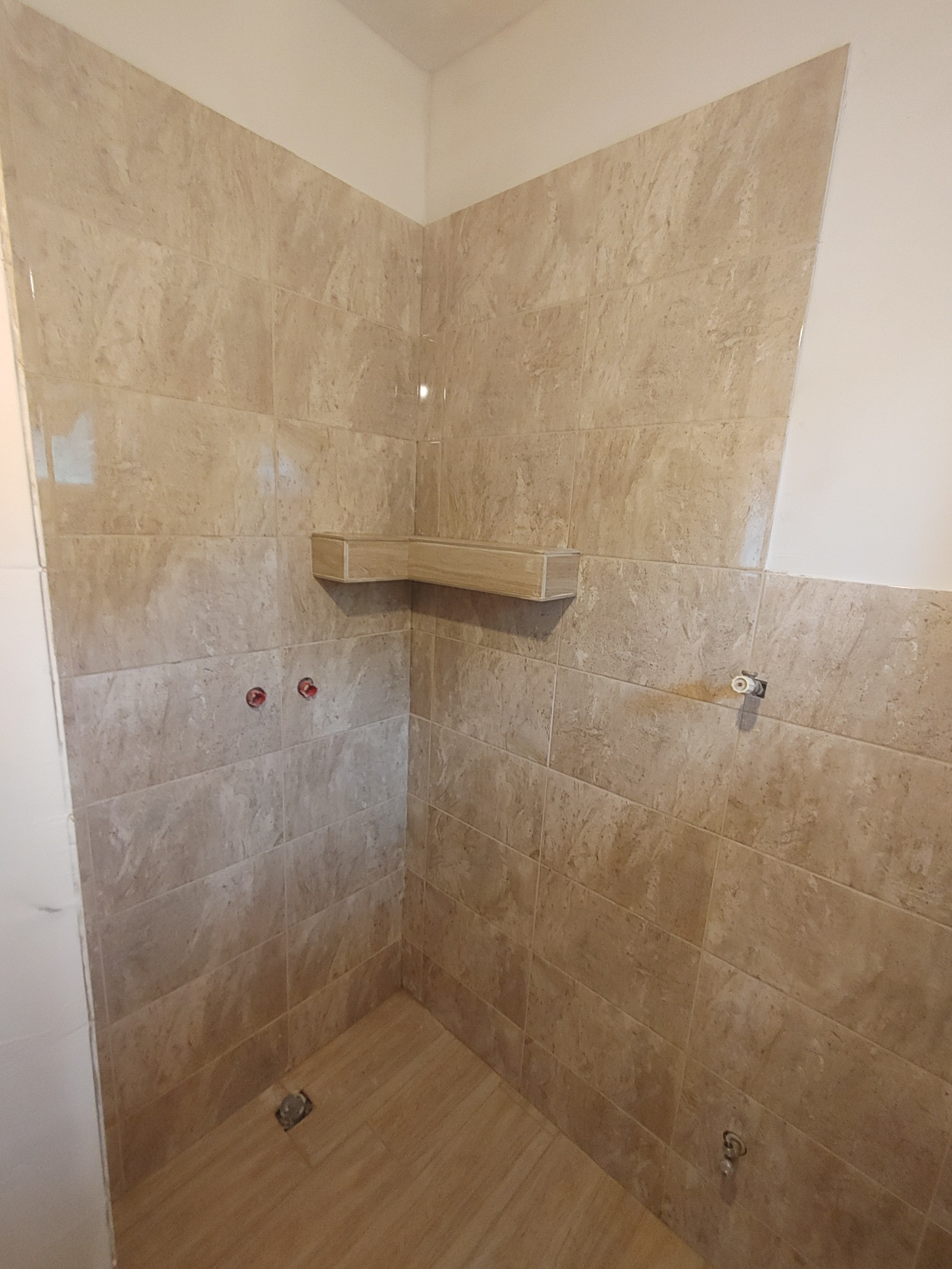 Wnętrze łazienki w trakcie prac wykończeniowych, widoczne beżowe płytki imitujące marmur na ścianach i podłodze, otwory na baterie i rury oraz półka z płytek.