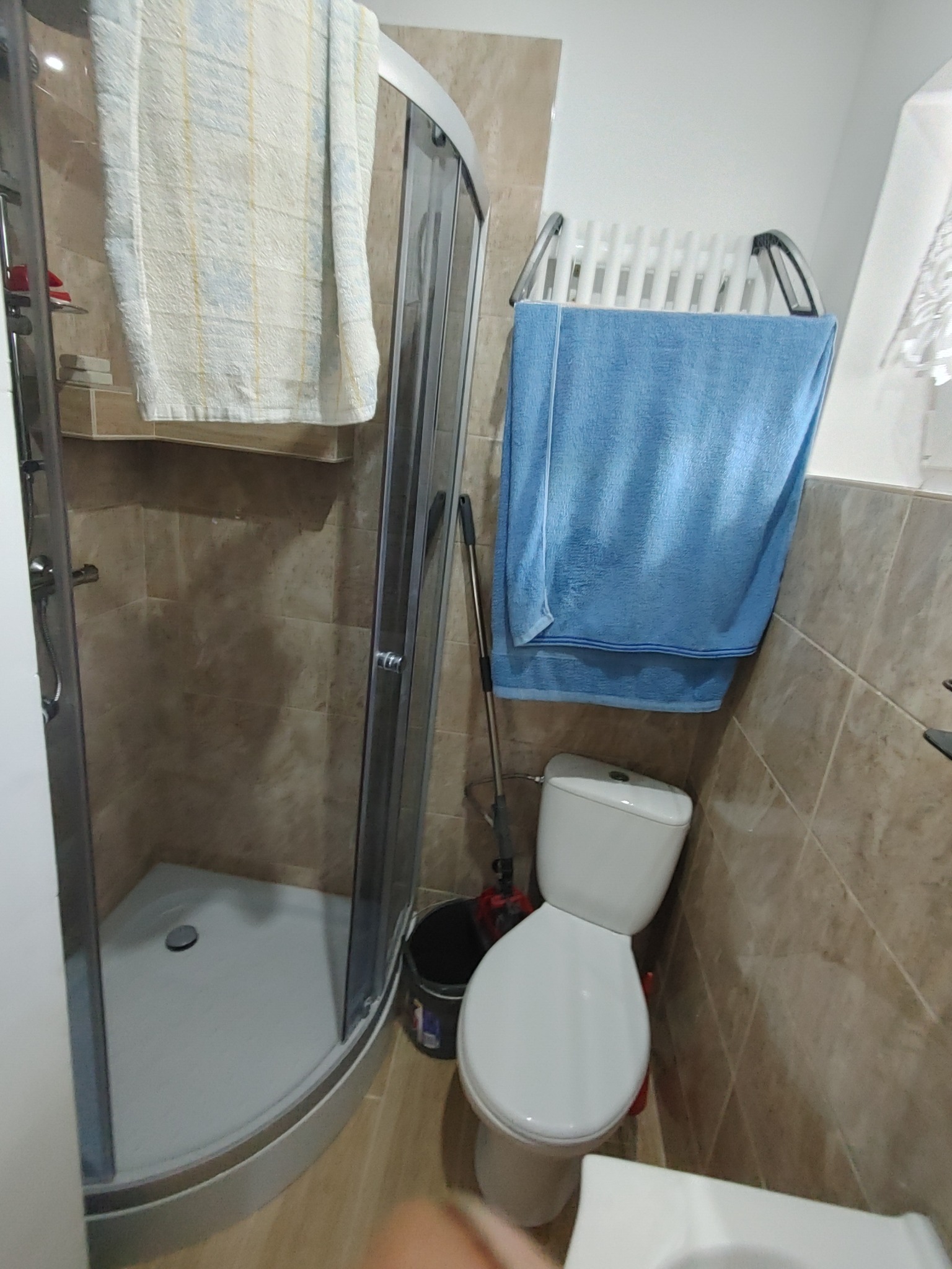 Narożna kabina prysznicowa z jasną zasłoną, biała toaleta z niebieskim ręcznikiem na grzejniku drabinkowym, beżowe płytki na ścianach, wiadro z mopem obok toalety.