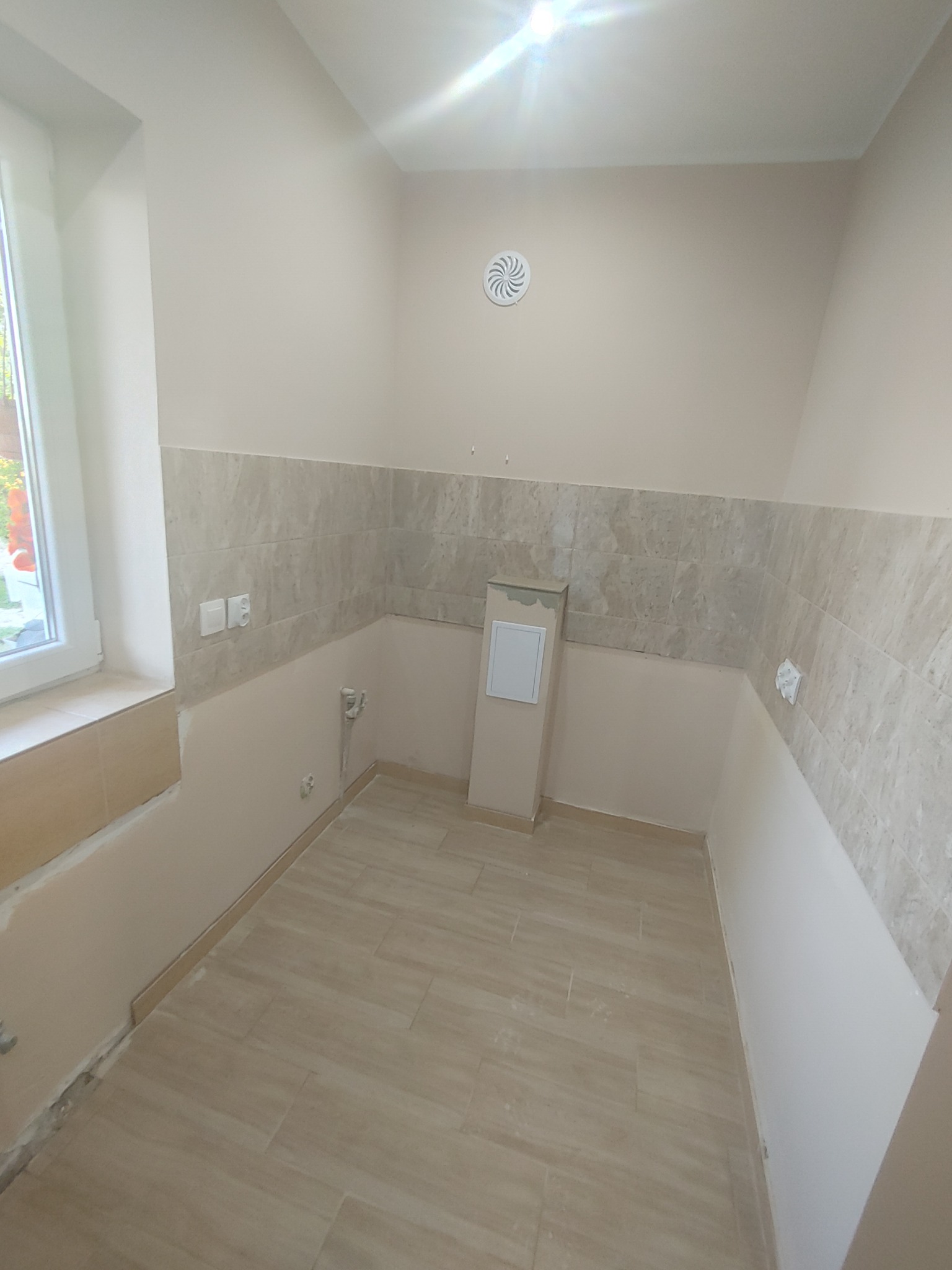 Wnętrze łazienki w trakcie układania płytek ceramicznych na podłodze i częściowo na ścianach, widoczna wentylacja sufitowa, okno z widokiem na zewnątrz, niedokończone prace wykończeniowe.