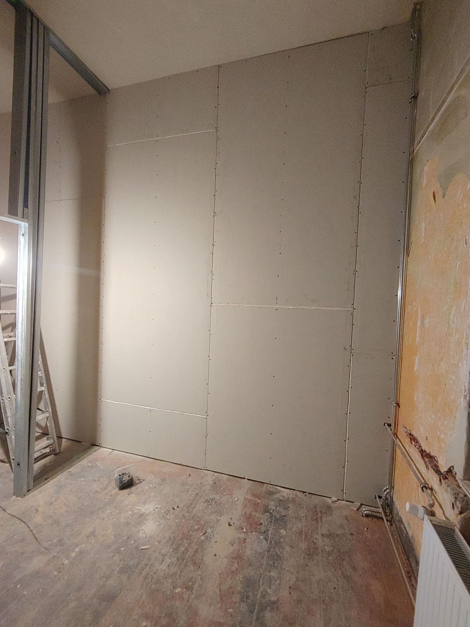 Montaż płyt gipsowo-kartonowych na ścianie w trakcie remontu mieszkania, widoczne profile metalowe, stara ściana z resztkami tapety i grzejnik.