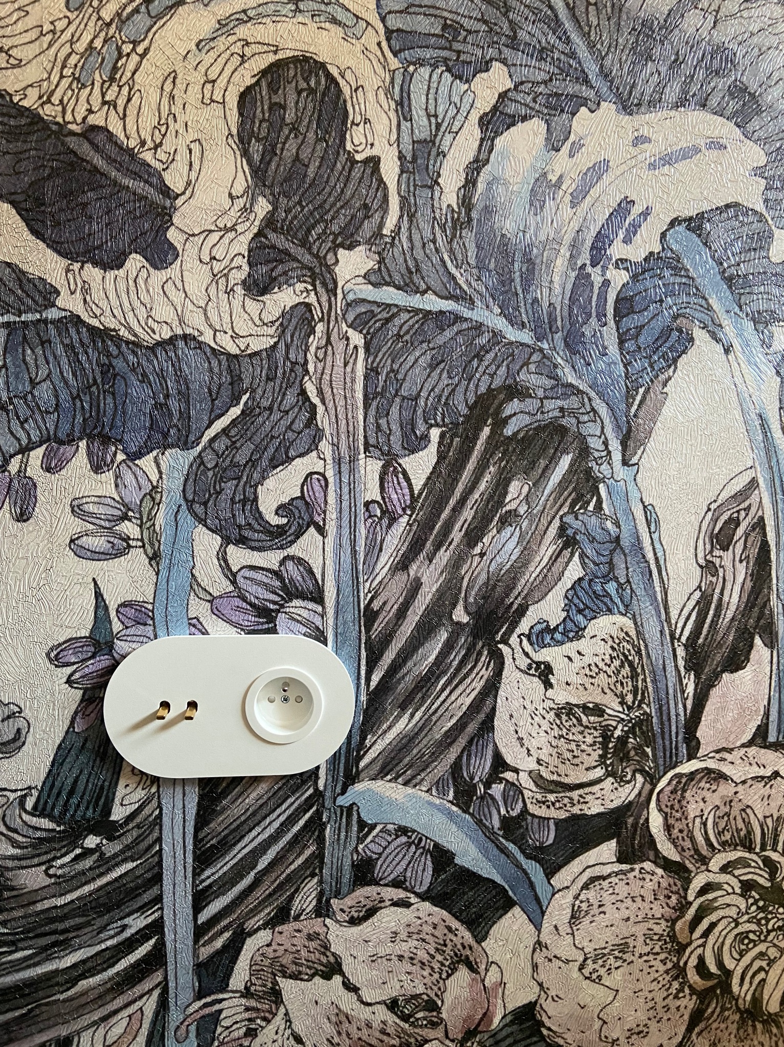 Białe gniazdko elektryczne z dwoma bolcami na tle ściany pokrytej tapetą z motywem kwiatowym w odcieniach bieli, szarości i fioletu.