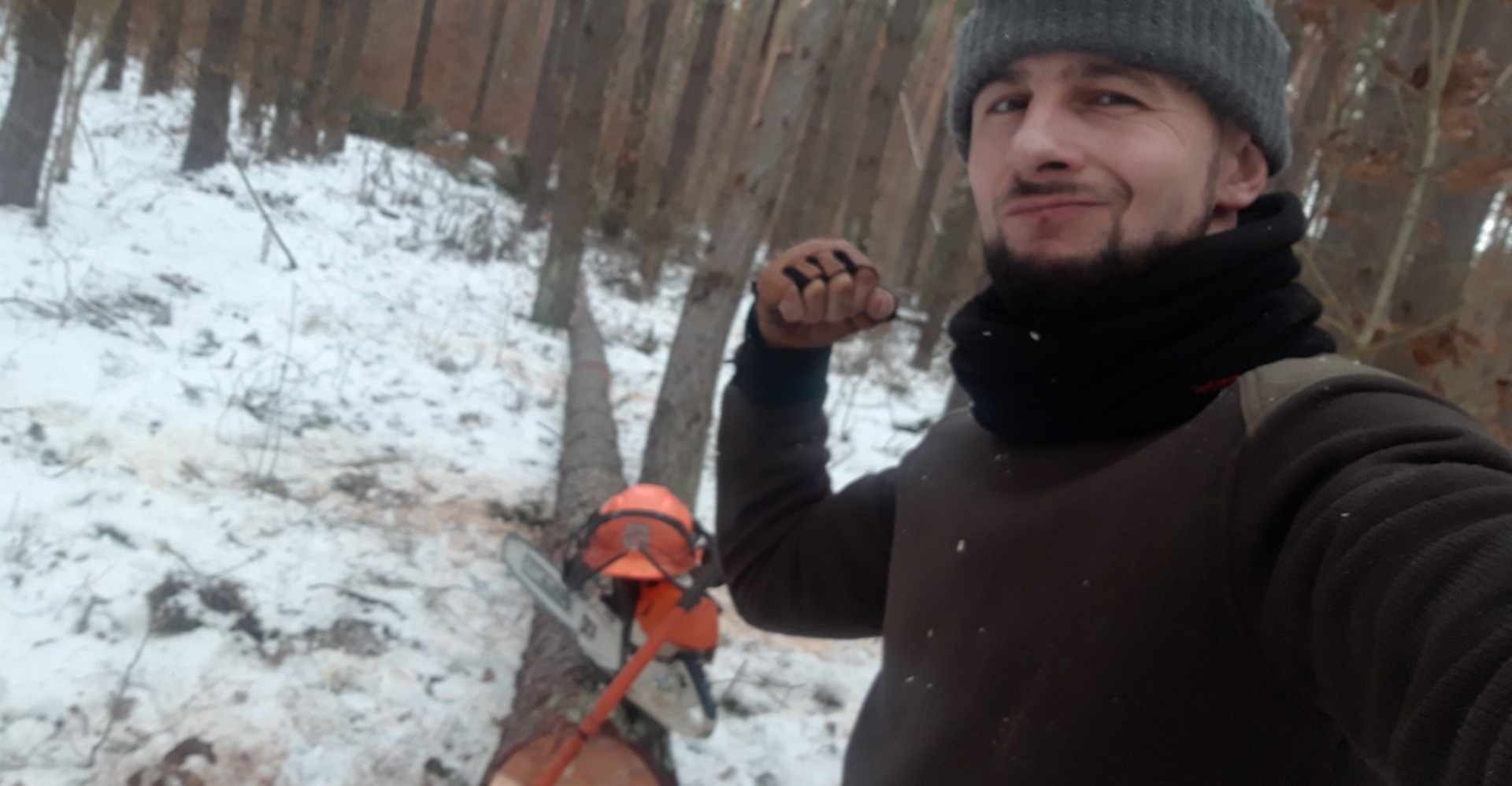 Mężczyzna w czapce i kominiarce robi selfie na tle ściętego drzewa w zimowym lesie. Na pniu leży kask ochronny i piła łańcuchowa. Mężczyzna unosi pięść w rękawicy z odkrytymi palcami.