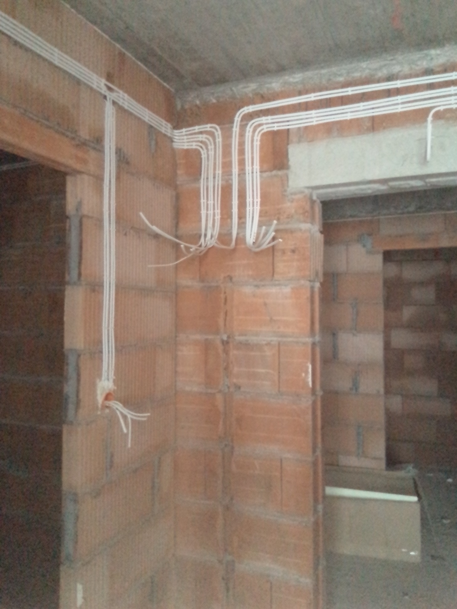 Surowa ściana z cegły z widocznymi białymi rurkami instalacji elektrycznej, wystające przewody z puszki w ścianie, widok na sąsiednie pomieszczenie.