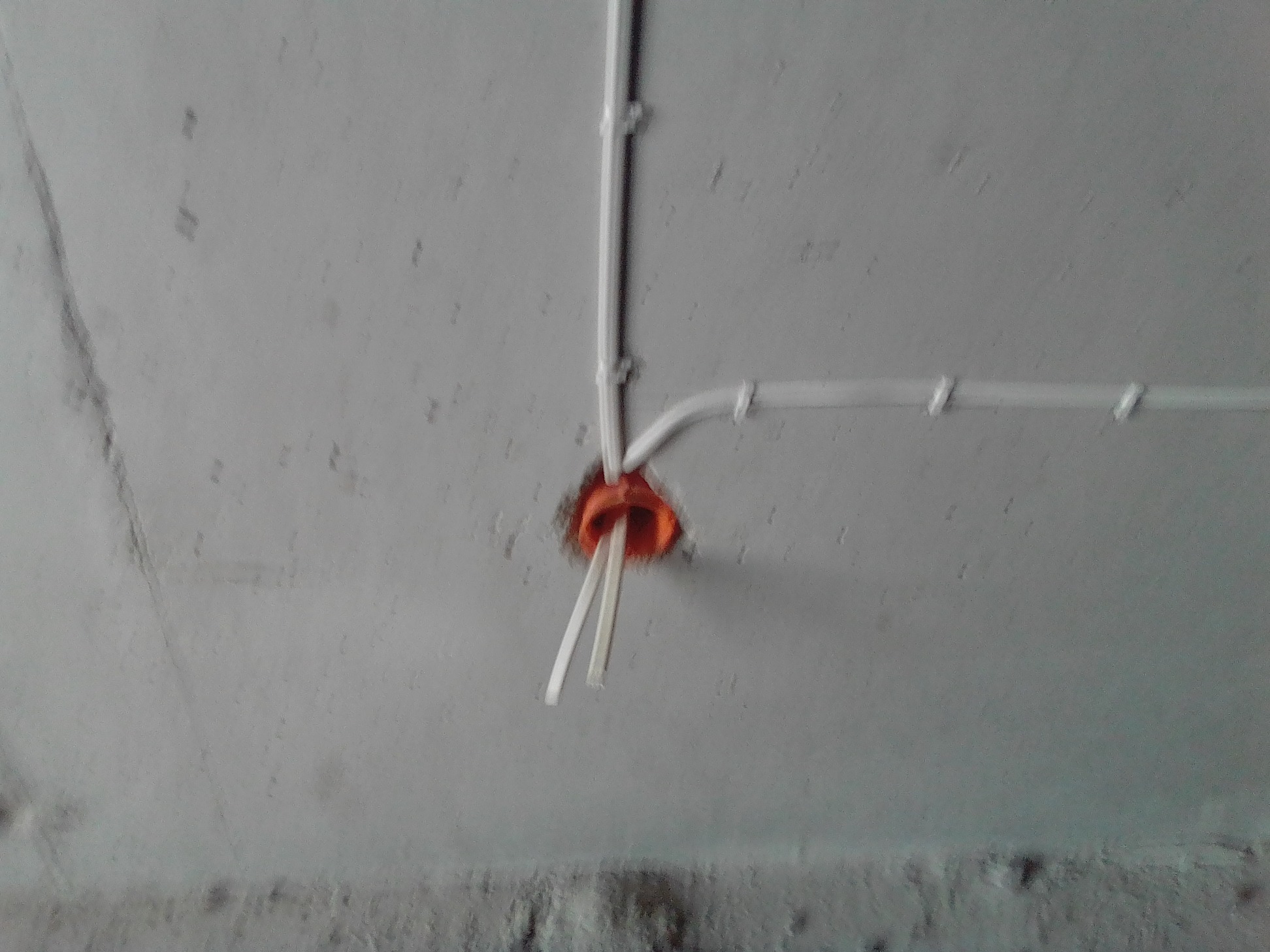 Białe przewody elektryczne wychodzące z pomarańczowej puszki instalacyjnej zamontowanej w szarym, surowym suficie.