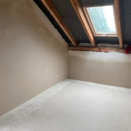 Poddasze po tynkowaniu maszynowym, widoczne drewniane elementy konstrukcji dachu, okno dachowe i fragment czerwonej tkaniny.