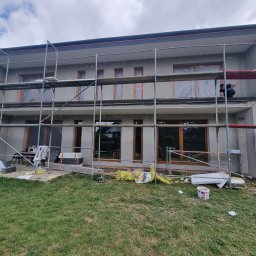 Maksteam Sp. z o.o. - Dwukondygnacyjny dom w trakcie ocieplania elewacji, z widocznymi rusztowaniami i pracownikiem na górnym poziomie. Okna zabezpieczone folią, materiały izolacyjne na trawniku.