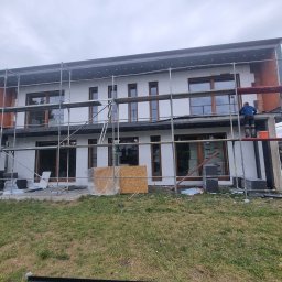 Maksteam Sp. z o.o. - Dwukondygnacyjny dom w trakcie ocieplania elewacji, widoczne rusztowania i pracownik na górze. Część ściany wykończona czerwoną cegłą, reszta w trakcie izolacji styropianem.