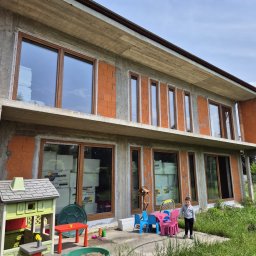 Maksteam Sp. z o.o. - Surowy stan dwukondygnacyjnego domu z betonu i cegły, z oknami w drewnianych ramach. Na tarasie dziecięce mebelki i bawiące się dziecko. W oknach widoczne materiały izolacyjne.