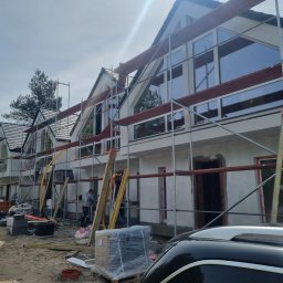 Maksteam Sp. z o.o. - Elewacja szeregowca w trakcie prac wykończeniowych, widoczne rusztowania i pracownik. Duże okna z widokiem na las. Częściowo widoczny samochód.