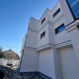 Maksteam Sp. z o.o. - Nowoczesny budynek z białą elewacją i ciemnymi obramowaniami okien, widziany z dołu pod kątem. Widoczne fragmenty ogrodu i niebieskie niebo.