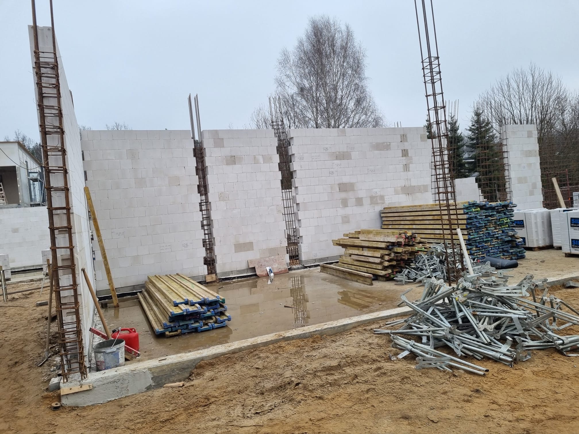 Wznoszenie ścian z bloczków betonowych z widocznym zbrojeniem pionowym. Na pierwszym planie stosy desek szalunkowych i metalowe podpory. Budowa w trakcie realizacji.