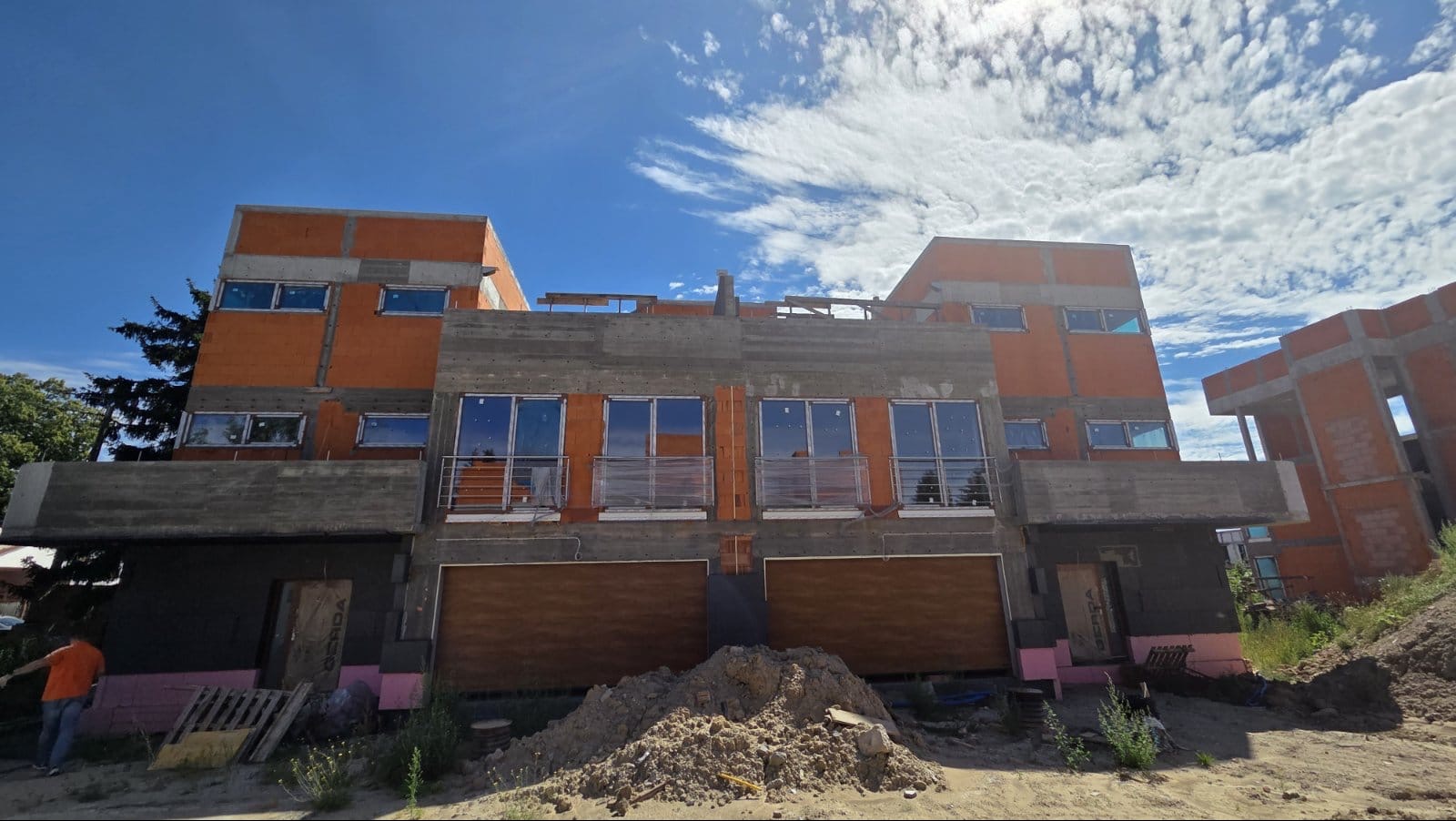 Wznoszone budynki w stanie surowym otwartym, z widoczną konstrukcją z betonu i cegły, okna wstawione, teren budowy z hałdą piasku na pierwszym planie.