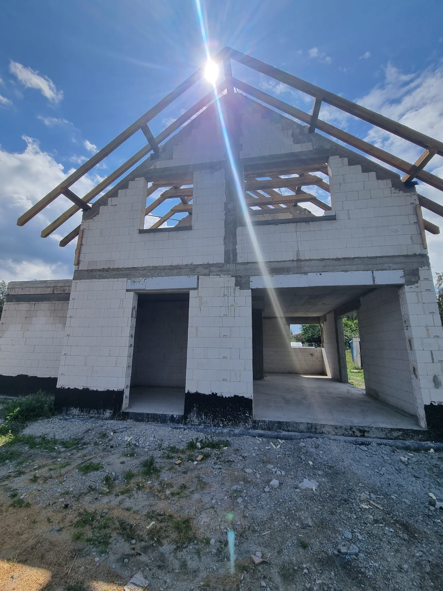 Surowy stan dwukondygnacyjnego domu z białego bloczka, widoczne otwory okienne i zarys dachu pod błękitnym niebem z promieniami słońca. Etap murowania ścian.