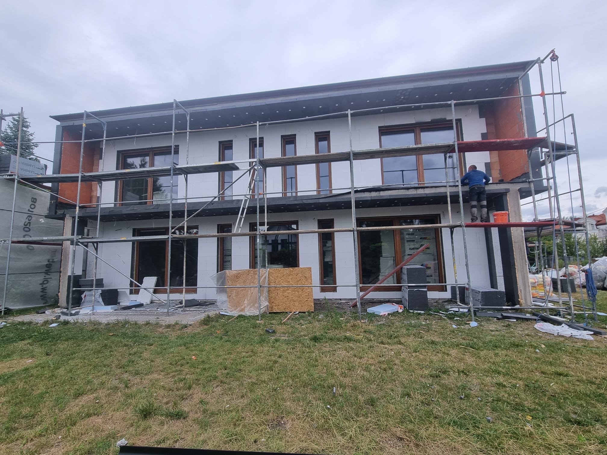 Dwukondygnacyjny dom w trakcie ocieplania elewacji, widoczne rusztowania i pracownik na górze. Część ściany wykończona czerwoną cegłą, reszta w trakcie izolacji styropianem.