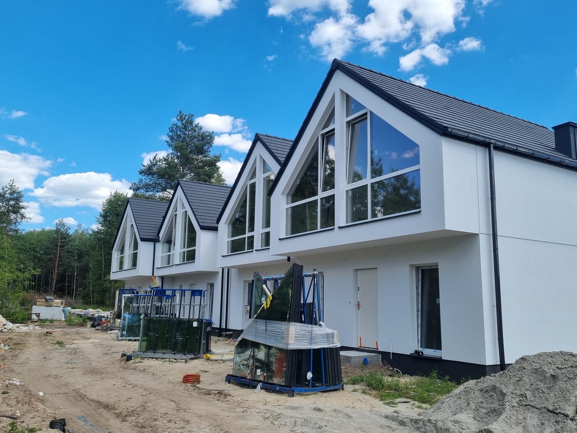 Nowoczesne, białe domy jednorodzinne z dużymi oknami, jeszcze przed montażem szyb. Na pierwszym planie przygotowane pakiety szklane. Błękitne niebo z chmurami w tle.