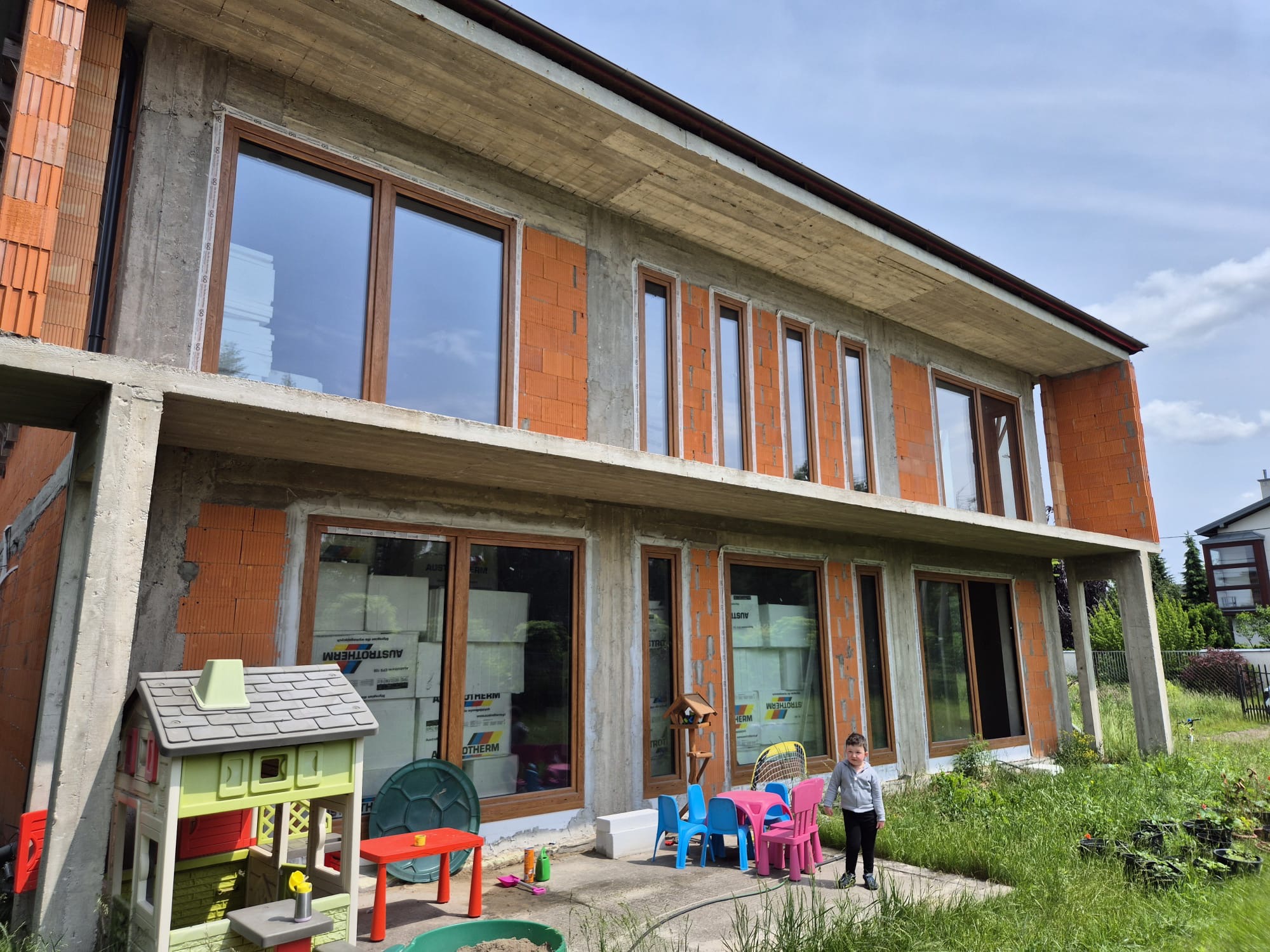 Surowy stan dwukondygnacyjnego domu z betonu i cegły, z oknami w drewnianych ramach. Na tarasie dziecięce mebelki i bawiące się dziecko. W oknach widoczne materiały izolacyjne.