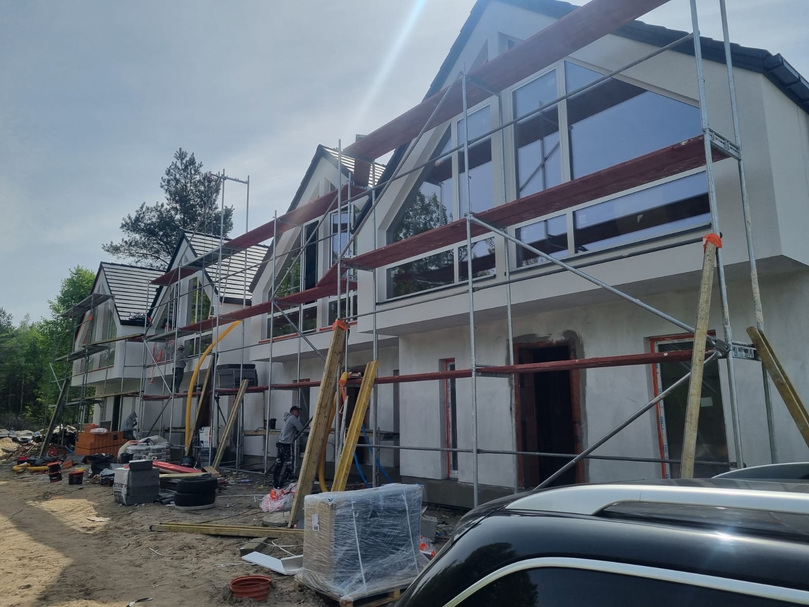 Elewacja szeregowca w trakcie prac wykończeniowych, widoczne rusztowania i pracownik. Duże okna z widokiem na las. Częściowo widoczny samochód.