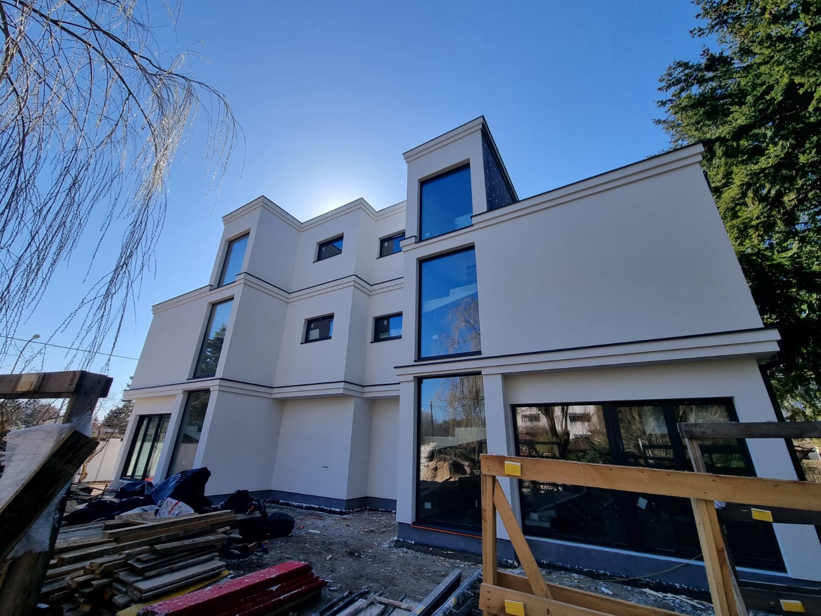 Nowoczesny, trzypiętrowy budynek z białymi ścianami i dużymi oknami w czarnych ramach, widziany z dołu pod kątem. Na pierwszym planie materiały budowlane i fragment drzewa.