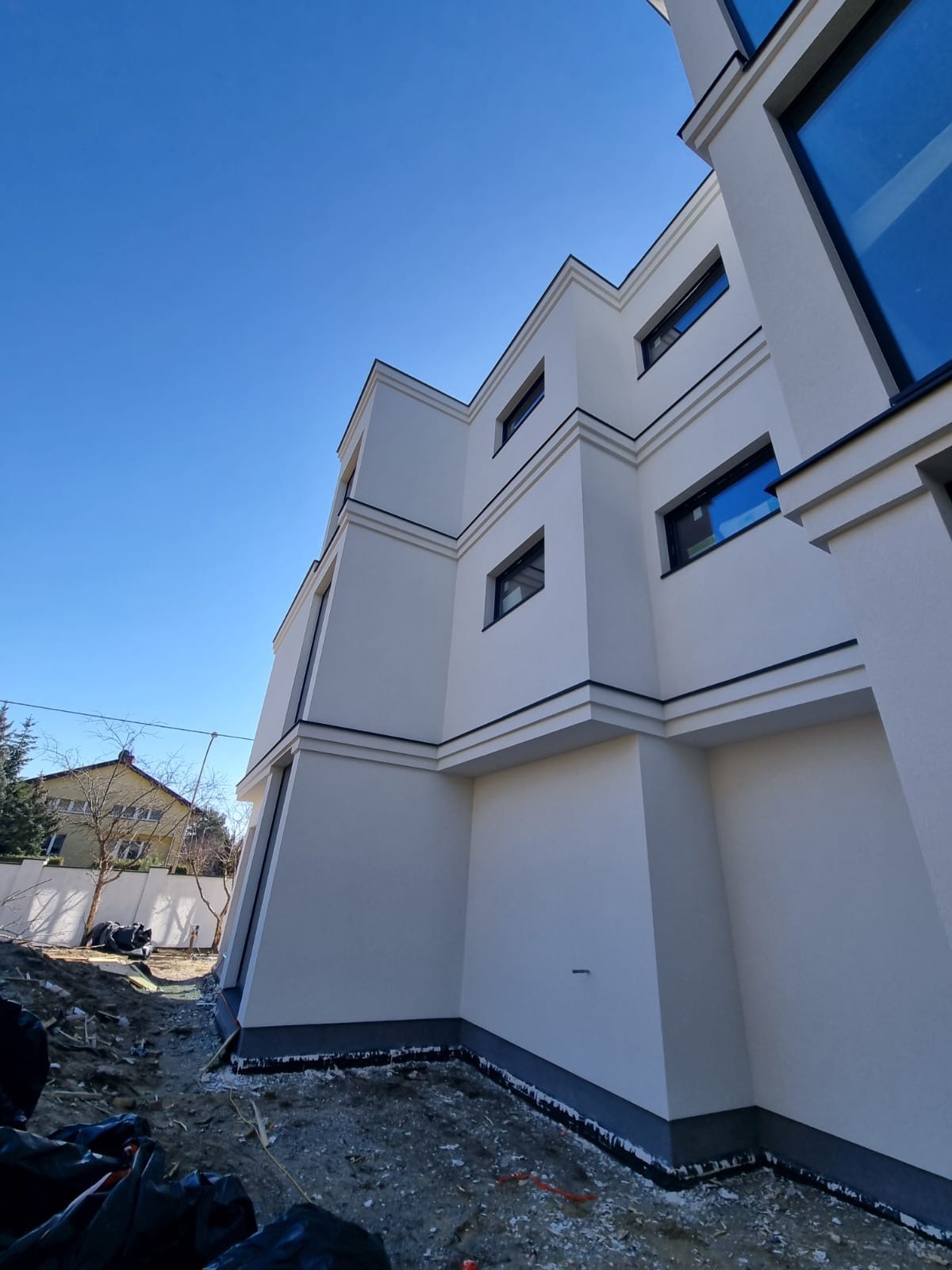 Nowoczesny budynek z białą elewacją i ciemnymi obramowaniami okien, widziany z dołu pod kątem. Widoczne fragmenty ogrodu i niebieskie niebo.