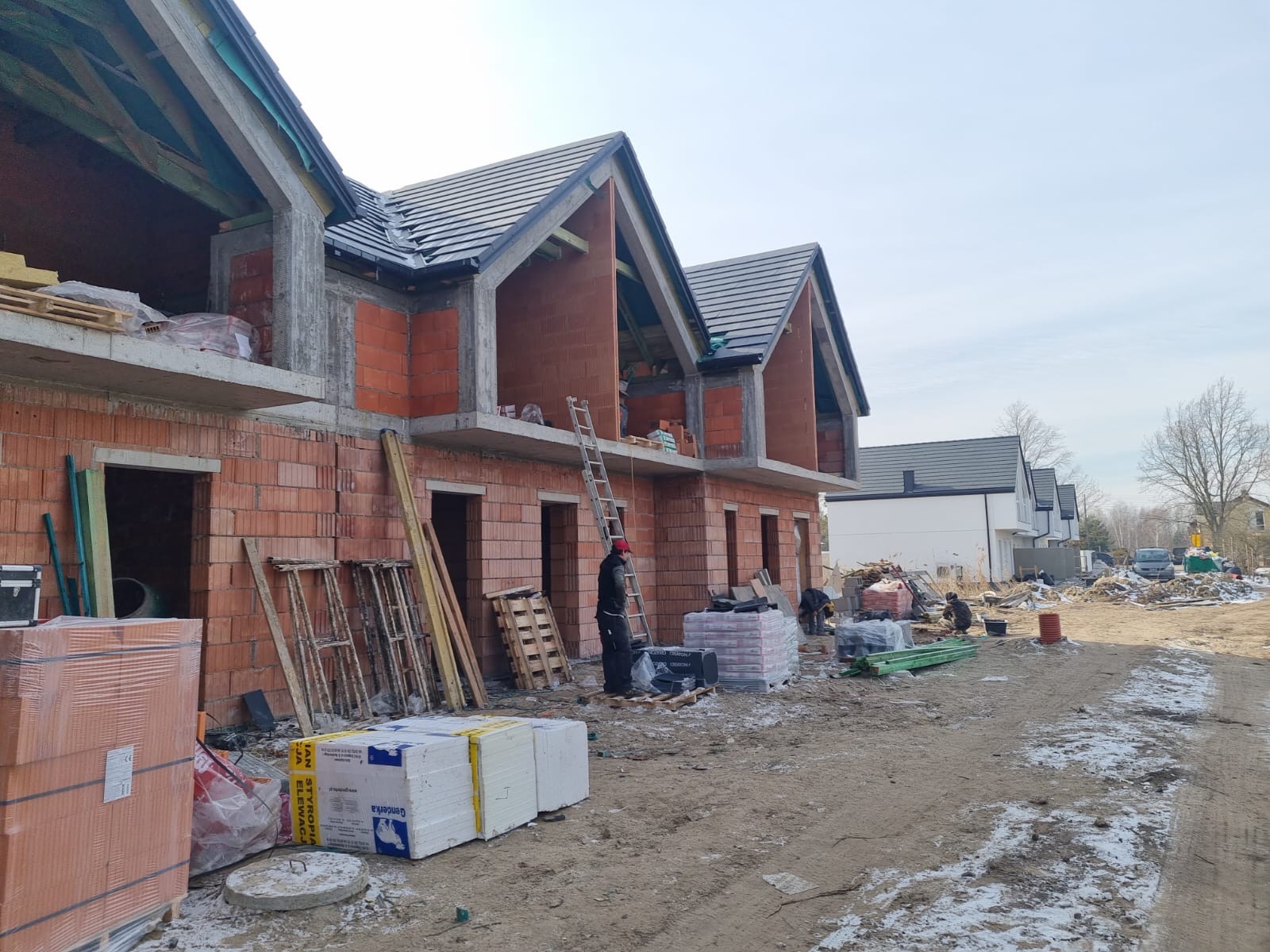Surowy stan budowy bliźniaka z czerwonej cegły, widoczne pustaki, betonowe stropy, drewniana konstrukcja dachu i materiały budowlane na placu. Częściowo ośnieżony teren.